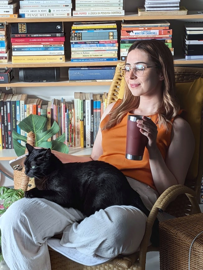 Vitória em sua poltrona de leitura, com estante de livros e seu gato preto